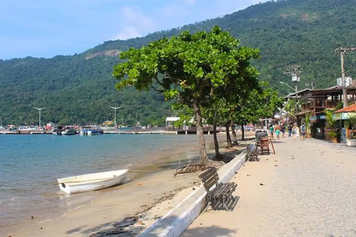 Paseo costero con árboles, bancos y un bote pequeño en la arena, con un muelle y el cerro verde al fondo