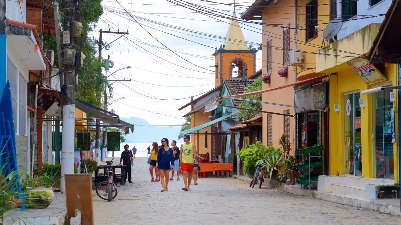 Rua de pedras com lojas coloridas e pessoas caminhando, com a torre de uma igreja e o mar ao fundo